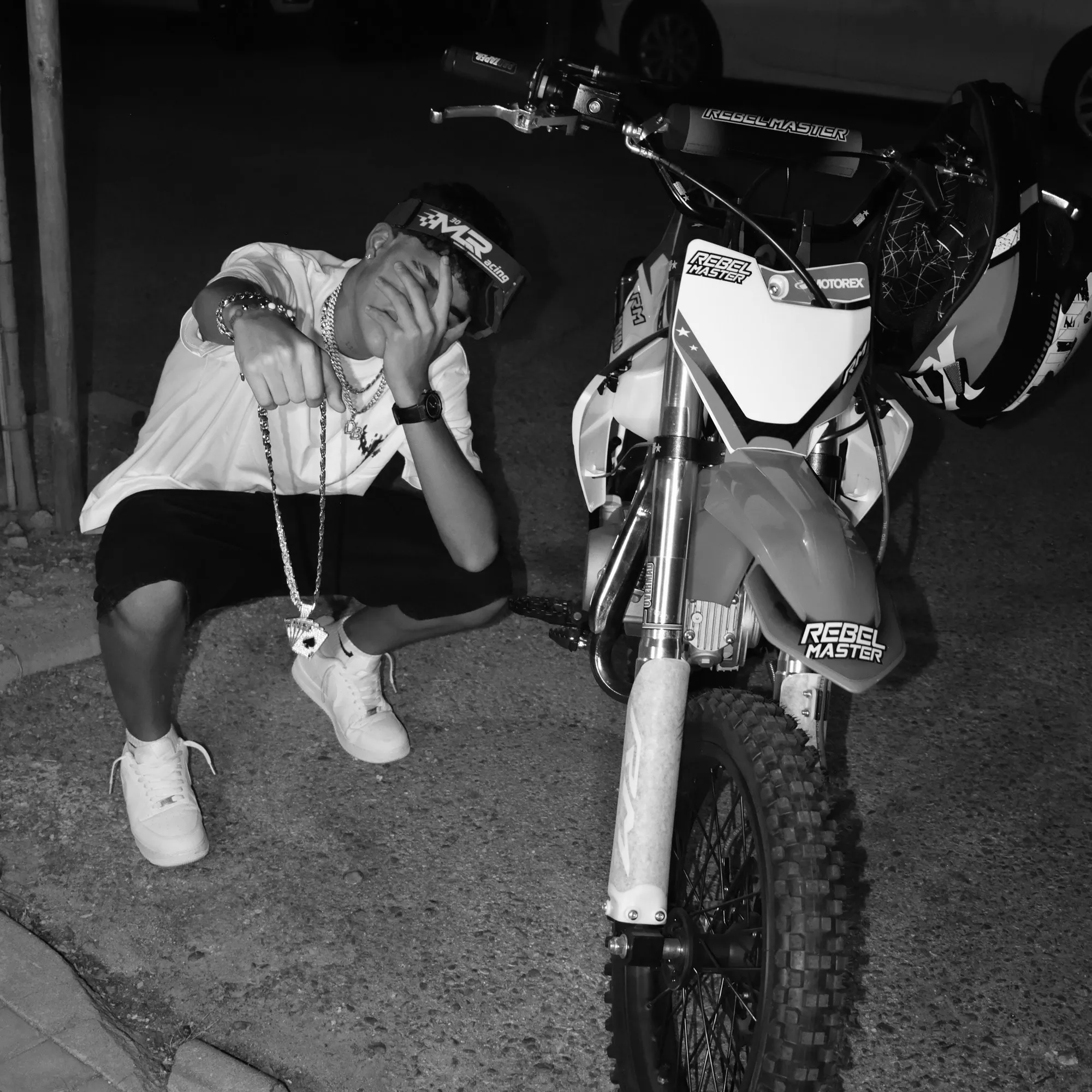 YOVNGBY agachado junto a una moto de motocross, de noche y con flash, tapándose la cara con la mano